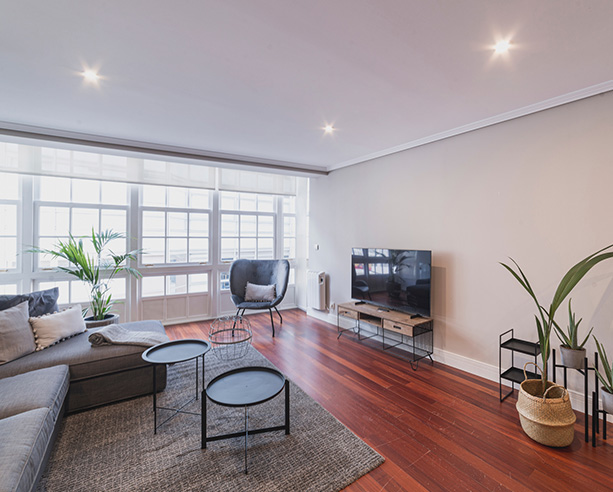 Salón luminoso con sofá en L, televisión y ventanales en apartamento de alquiler en A Coruña